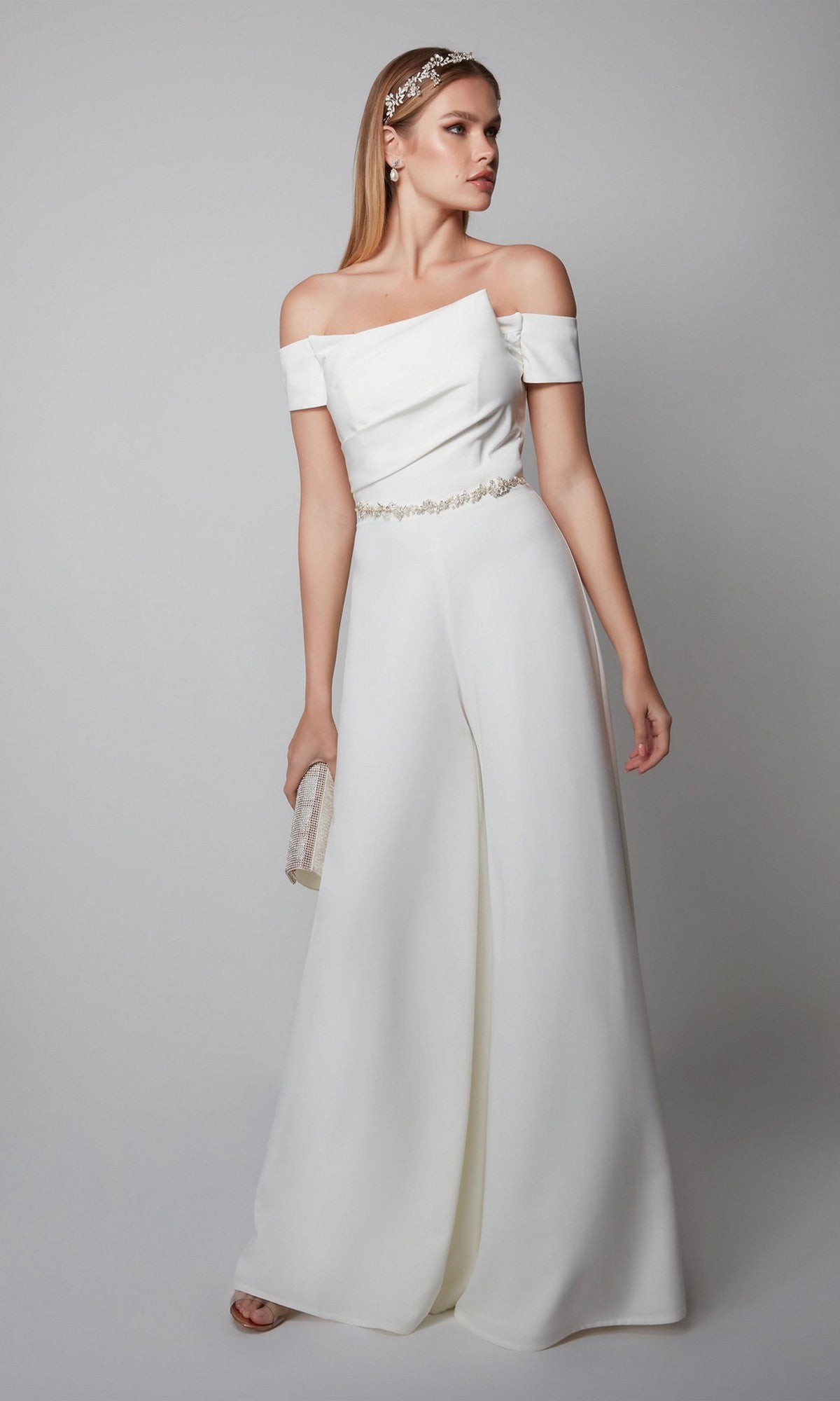 Woman wearing the Long Formal Jumpsuit: Alyce 70014, featuring an off-the-shoulder design and wide legs, paired with a small clutch and jeweled headband and earrings against a plain background.